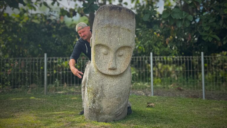 Mężczyzna z czapką z daszkiem za kamiennym posągiem z trzema palcami na dłoni, na tle zieleni i ogrodzenia
